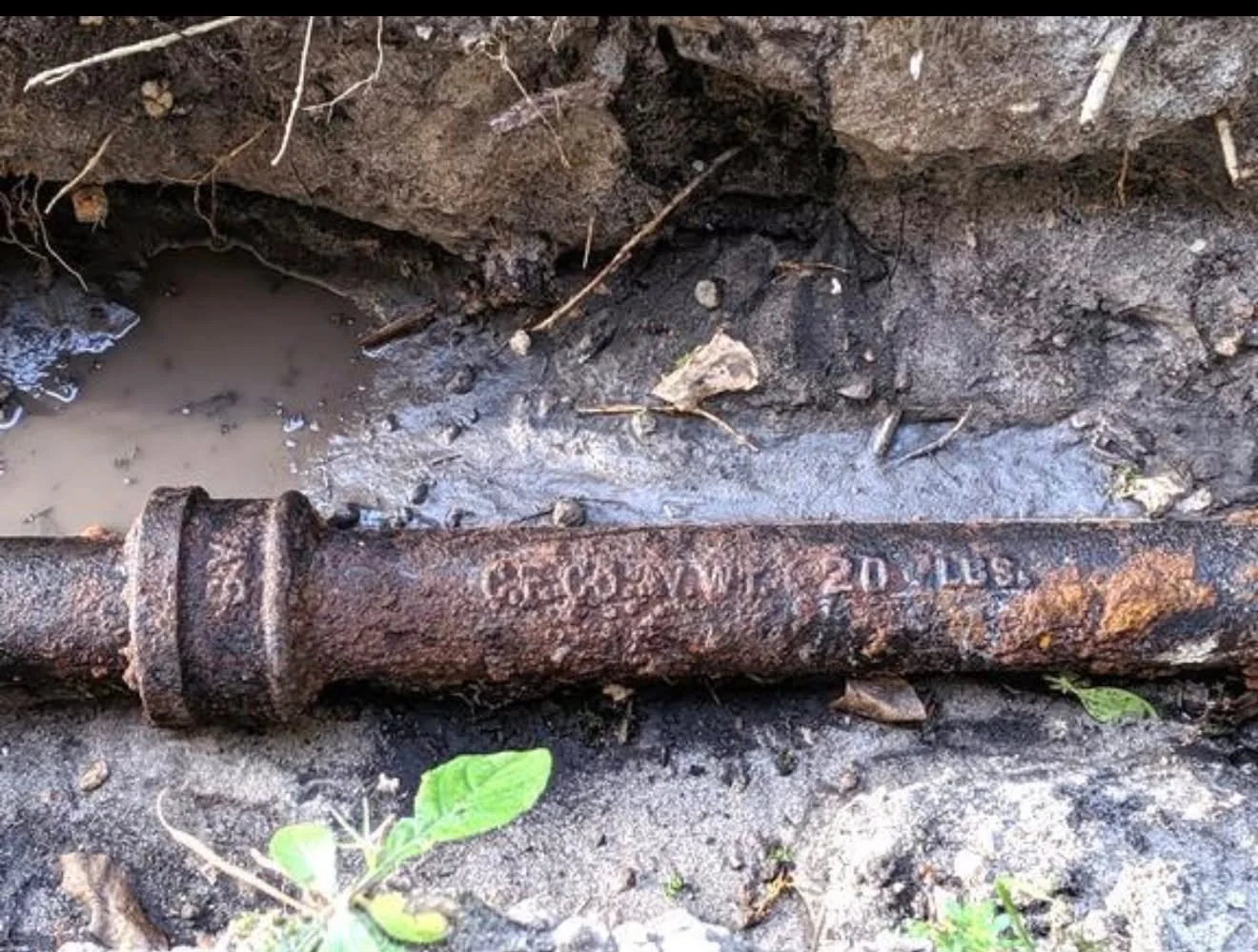 A badly corroded cast iron sewer pipe lying in an open trench, the wall flaking away