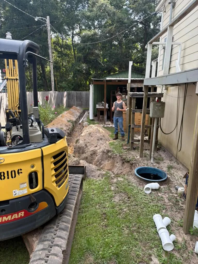 A yellow mini excavator digging a trench alongside a house with pipe staged beside it