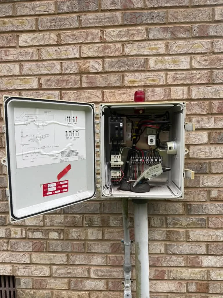 A grinder pump control panel mounted on a post against brick, cover open to the controls
