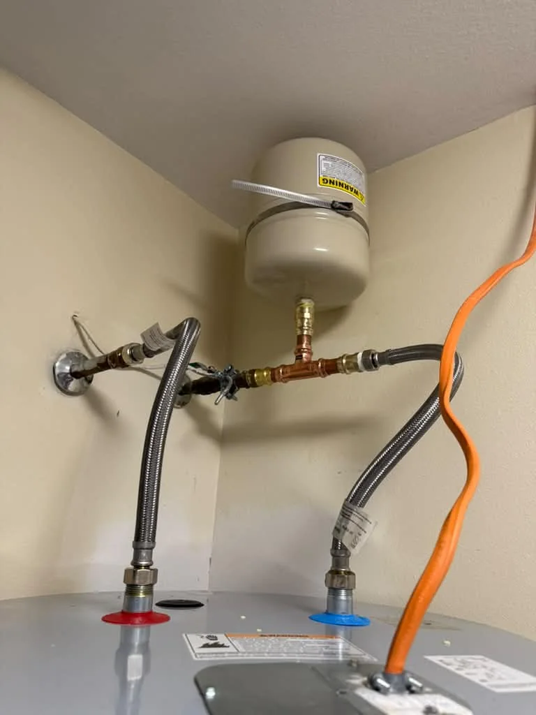 A water heater top with braided connectors, an expansion tank and an orange condensate hose in an attic space