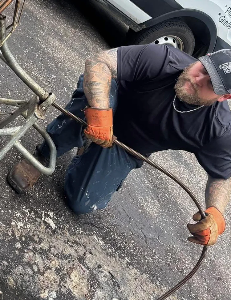 Paul Evans kneeling on a driveway in orange work gloves, feeding a drain machine cable, the LT Drain van behind him