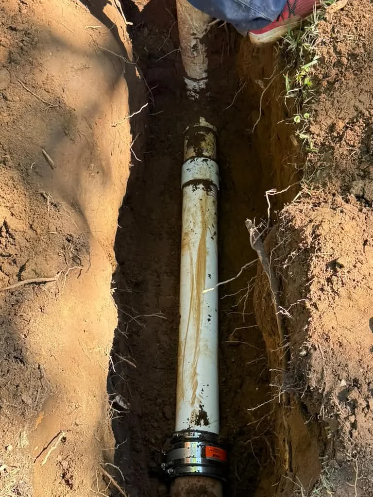 A deep narrow trench exposing a white PVC sewer line with a coupling, red clay walls on both sides