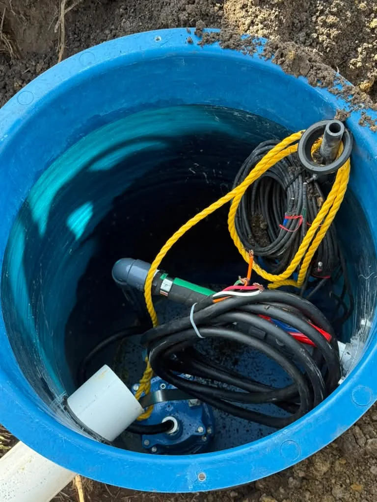 Looking down into a blue grinder pump basin set in the dirt, pump, lift rope and coiled cables inside
