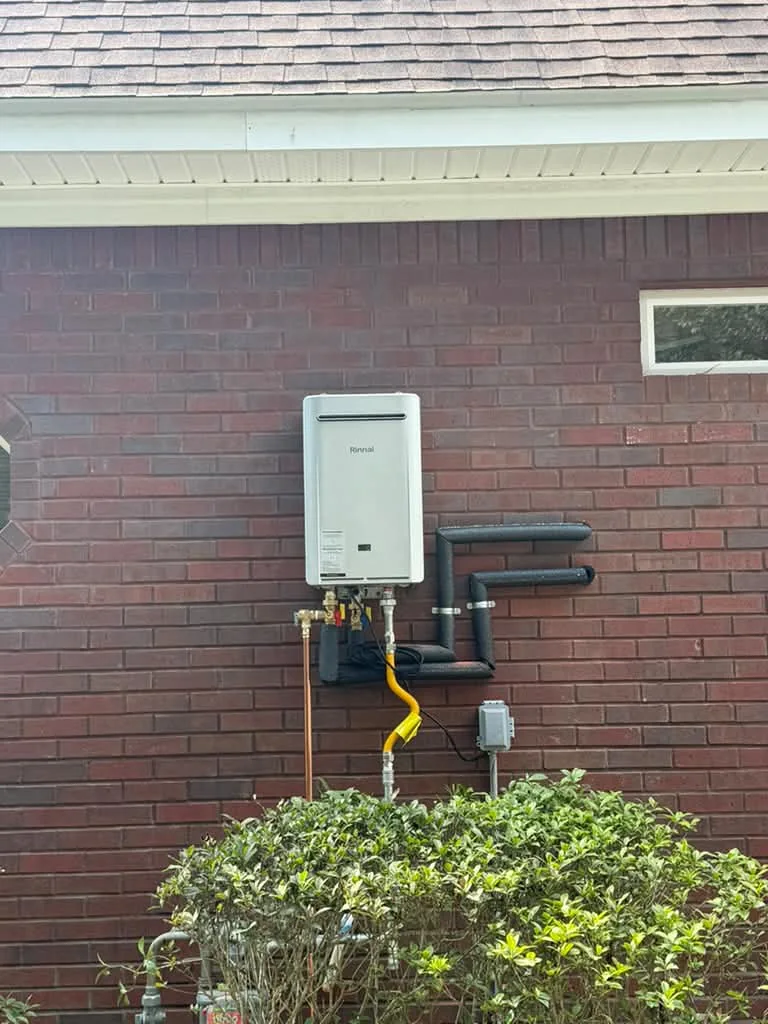 A tankless water heater mounted outdoors on brick above a shrub, yellow gas line and copper connections